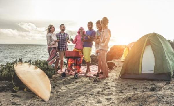 Happy friends drinking beers at camping barbecue picnic next to the ocean - Surfers people havin<em></em>g fun and laughing together - Main focus on right guys - Travel, vacation and friendship concept