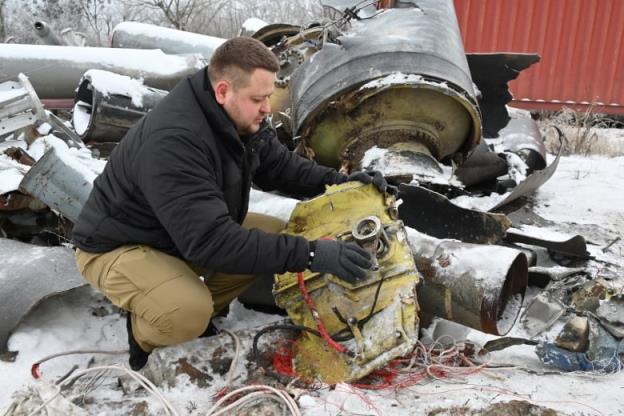 A bearded man wearing gloves crouches on a snowy filed to hold and examine a large piece of me<em></em>tal that co<em></em>ntains wiring.