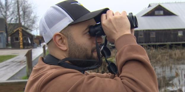 A man in a ba<em></em>seball cap is shown in profile holding binoculars to his eyes