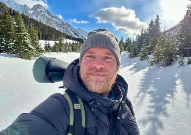 A man takes a selfie in front of snowy mountains. 