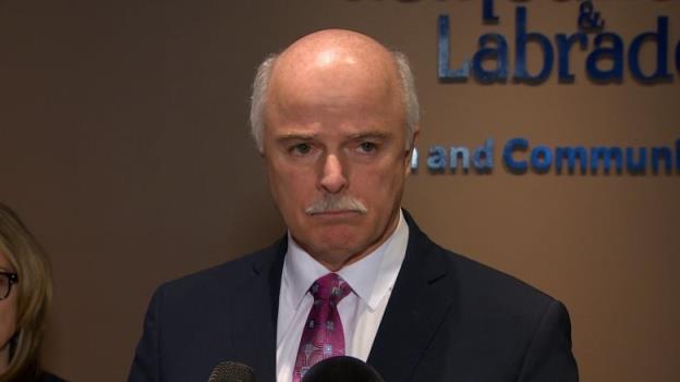 A man wearing a suit stands in front of a beige wall.