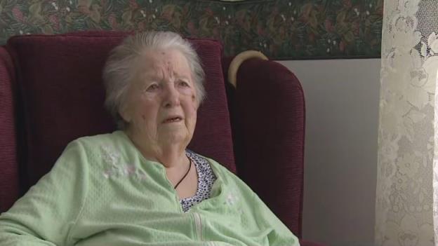 An elderly white woman with white hair sits in a maroon armchair. She's wearing a green blouse.