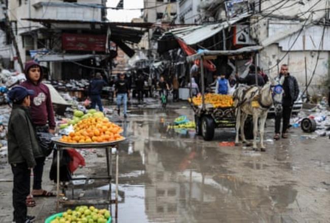 People stand on a wet and muddy road selling fruit from a donkey-led cart and a simple stand