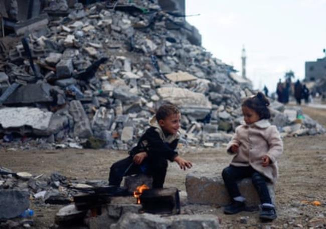 Two small children sit next to a small fire on a street surrounded by rubble