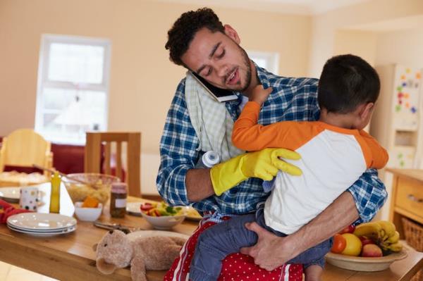 A man talks into his cellphone while holding a child. He has a cleaning glove on one hand and there's a baby bottle under his arm.