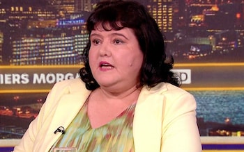 Ms Harvey wears a light-yellow jacket and sits in front of a video backdrop of a city at night
