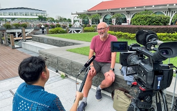 A man sits on a low wall in shorts being filmed and interviewed by a man holding a microphone marked 'Reuters'