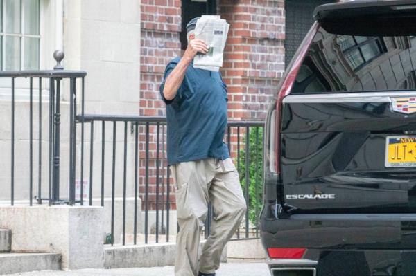 Robert De Niro holds a newspaper to his face as he walks out of building entrance. 