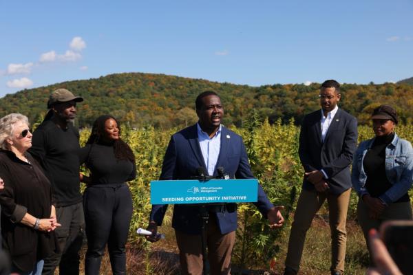 Chris Alexaner speaking at a podium in front of a marijuana field on a blue sky day with five other people to his side. 