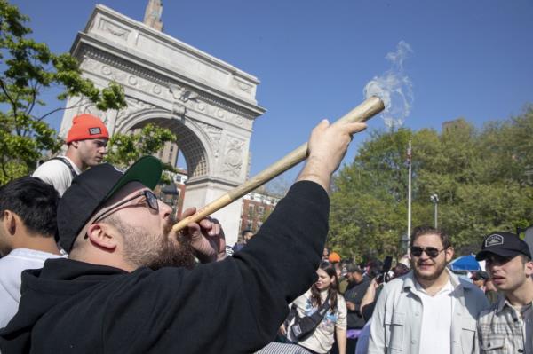 A bearded man smoking a three foot long blunt in washington square park with a crowd of people watching. 