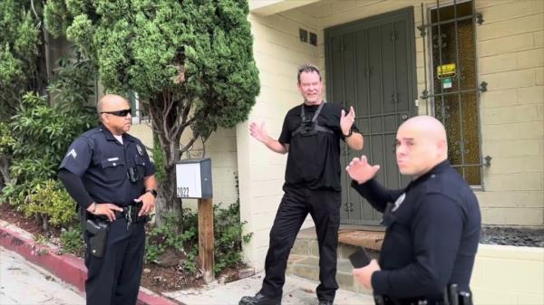 Flash Shelton is dressed in black and joined by two police officers while standing outside a home.
