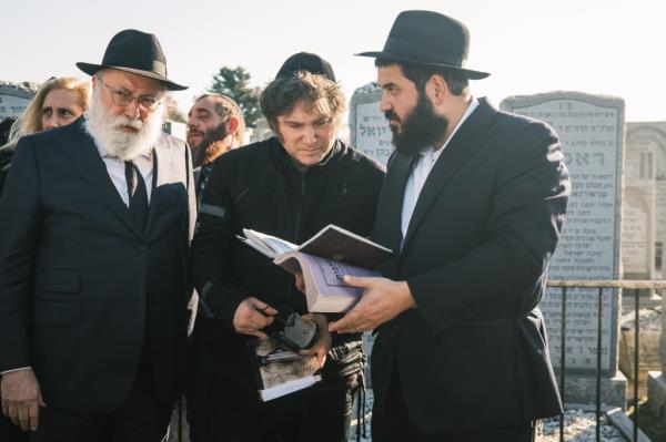 Three men, one holding an open book, stand together in a graveyard in Queens.