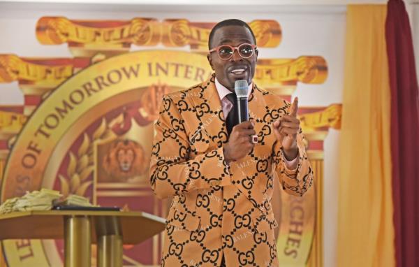 Lamor Whitehead in an orange Balenciaga suit speaks into a microphone during a sermon.  