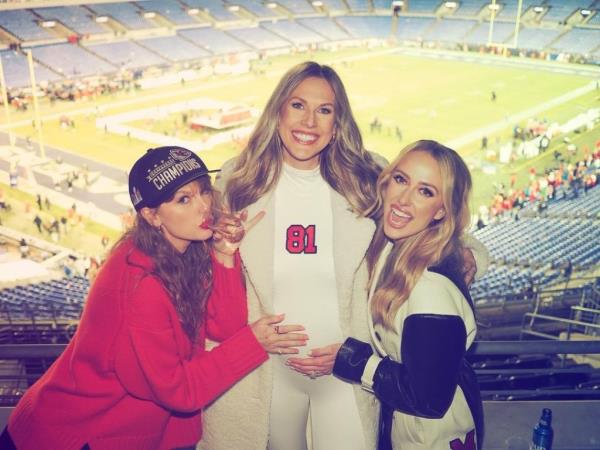 Taylor Swift, Lindsay Bell and Brittany Mahomes wearing AFC Champio<em></em>nship hats posing for a picture with smiles on their faces.