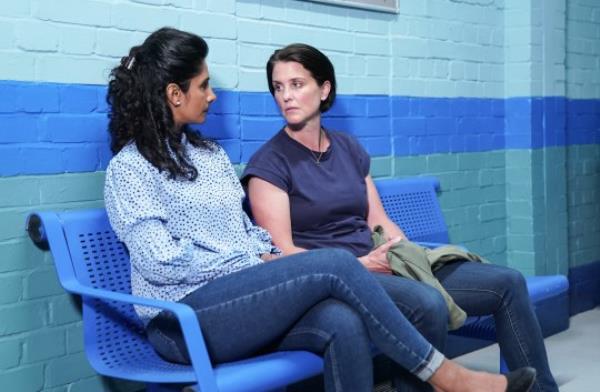 Suki and Eve look at each other while sitting in the corridor at the police station.