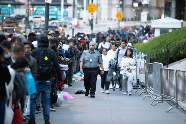A security officer leads migrants with women and children  on a sidewalk outside 26 Federal Plaza 