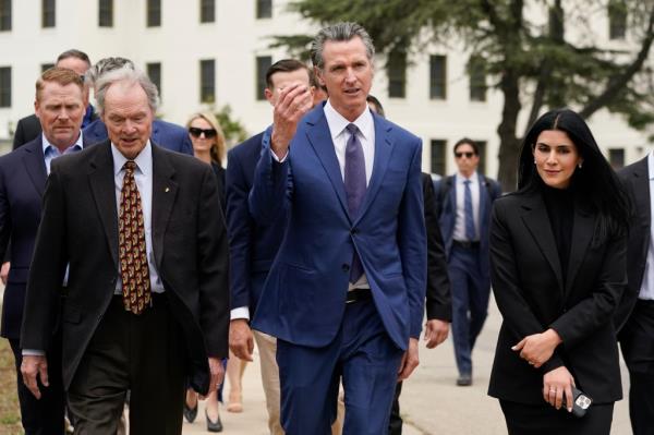 Gavin Newsom walking outside with an entourage around him as he gestures. 