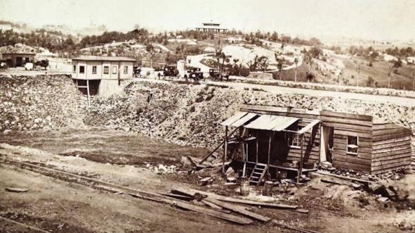 A photograph of a wooden home surrounded by a field, embankment and other homes in the former Seneca Village.