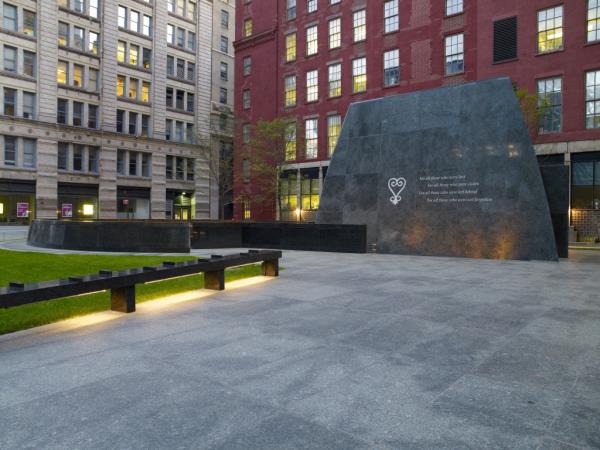 A photo of the modern day memorial at the African Burial Ground with slate squares on ground alo<em></em>ngside some grass with tall building on the sides.