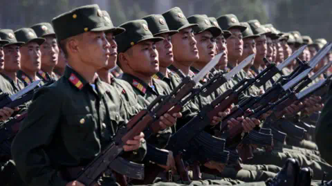 Getty Images Korean People's Army soldiers march during a mass rally on Kim Il Sung square in Pyongyang. They are seen marching in a line, each staring forward holding a rifle at a 45-degree angle. The rifles have bayo<em></em>nets on the end. 